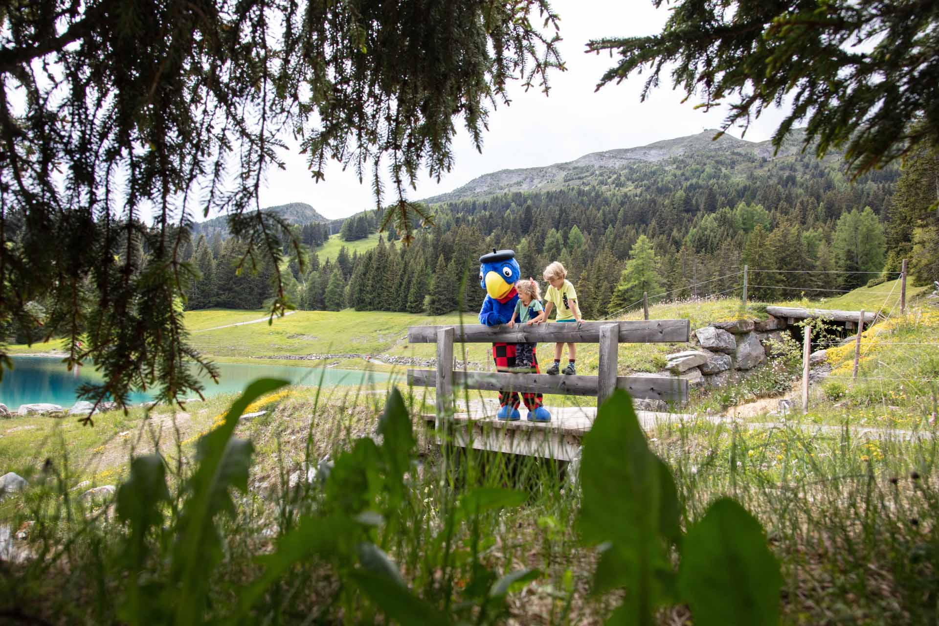 Zwei Kinder unterhalten sich mit Globi an einem Holzgeländer, im Hintergrund ist ein idylischer Bergsee